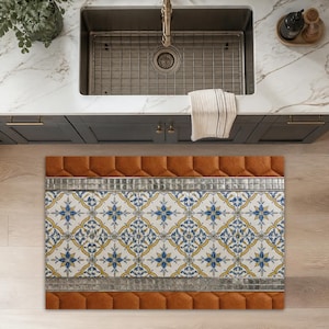 May include: A rectangular floor mat featuring a central panel with a repeating blue and yellow floral tile pattern, bordered by metallic silver mosaic strips and solid burnt orange quilted edges, positioned on a light wood floor in a kitchen.