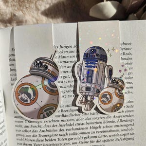 Könnte beinhalten: Drei Star Wars-Charakteraufkleber: BB-8, R2-D2 und ein kleinerer BB-8. Die Aufkleber sind weiß mit orangefarbenen, blauen und silbernen Details. Sie befinden sich auf einem Buch mit deutschem Text.