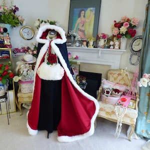 May include: A floor-length hooded cloak featuring a deep black exterior and a vibrant red interior lining. The garment is trimmed with thick white faux fur along the hood and front edges, and it is paired with a matching white faux fur muff.