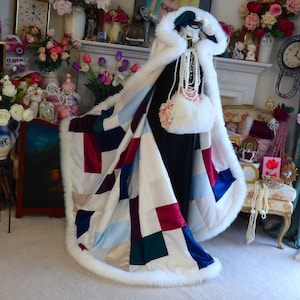 May include: A white faux fur hooded cape with a patchwork design featuring squares of burgundy, navy blue, light blue, green, and beige satin. The cape has a white faux fur trim around the edges.
