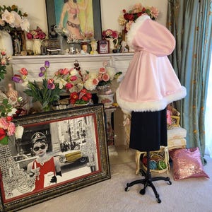 May include: A light pink hooded cape featuring a thick white faux fur trim along the hood and the bottom hem. The cape is displayed on a black mannequin stand, showcasing a flared silhouette and a smooth, solid-colored fabric construction.