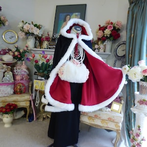 May include: A floor-length hooded cloak featuring a black exterior and a deep red interior lining. The garment is trimmed with thick white faux fur along the hood and front edges. A white faux fur muff with floral accents is attached to the front.