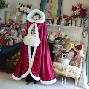 May include: A floor-length, deep burgundy satin cloak featuring a plush white faux fur trim along the hood, front opening, and hem. The garment includes a matching white faux fur muff accessory with small pink floral accents attached at the front.
