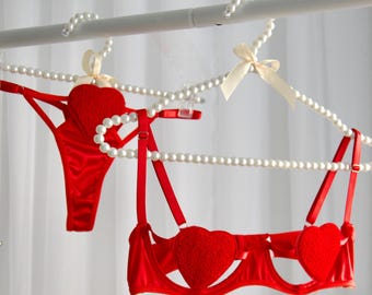 May include: Red lingerie set featuring a bra and thong, both adorned with heart-shaped accents. The bra has a unique cut-out design. The set is displayed on pearl-beaded hangers with cream-colored bows, against a white backdrop.