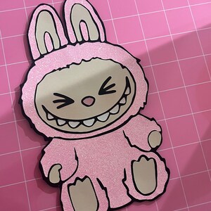 May include: Pink glitter bunny rabbit with a toothy grin and black eyes. The bunny is wearing a pink glitter costume with black outlines.