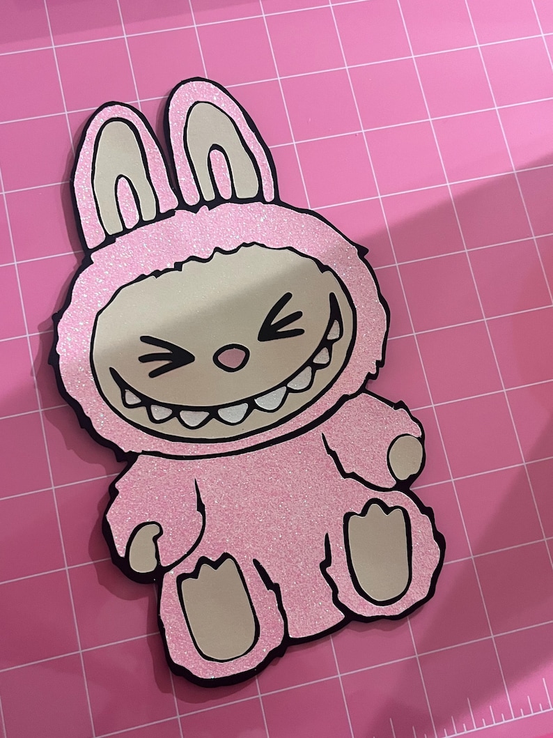 May include: Pink glitter bunny rabbit with a toothy grin and black eyes. The bunny is wearing a pink glitter costume with black outlines.