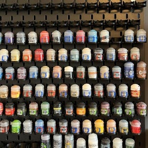 May include: A black wall-mounted pegboard organizer holding numerous small Citadel paint pots in various colors. The pots are arranged in horizontal rows within individual circular slots, featuring labels with the Citadel brand name.