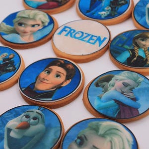 Peut inclure: Un ensemble de biscuits ronds décorés représentant des personnages du film d'animation La Reine des neiges. Les biscuits sont décorés avec des images d'Elsa, d'Anna, d'Olaf, de Kristoff et de Hans. Un biscuit porte l'inscription "FROZEN".