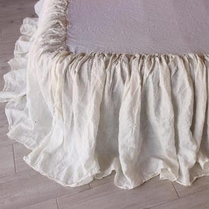 May include: A close-up of a bed with a white linen bed skirt. The bed skirt has a ruffled design and hangs down to the floor. The bed is covered with a white sheet.