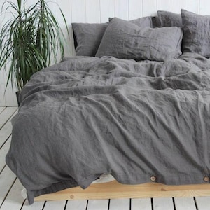 Könnte beinhalten: Ein Bett mit grauer Leinenbettwäsche, einschließlich Bettbezug und Kissenbezügen. Der Bettbezug hat Holzknöpfe am unteren Rand. Das Bett steht auf einem Holzrahmen, im Hintergrund eine grüne Pflanze.