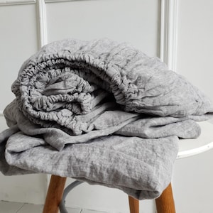 May include: A gray linen fitted sheet set, neatly folded on a small white table with wooden legs. The elasticized edge of the fitted sheet is visible, along with the soft, textured fabric of the sheets. The background is a white wall.
