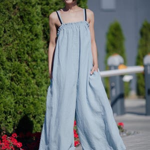 Könnte beinhalten: Eine Frau trägt einen hellblauen Leinen-Jumpsuit mit geraffter Taille und weiten Beinen. Sie lächelt und steht auf einem Bürgersteig mit grünen Büschen im Hintergrund.