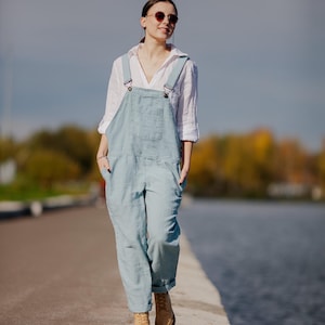 Könnte beinhalten: Eine Frau in einem weißen Hemd und einer hellblauen Jeanslatzhosen geht auf einem Betonweg entlang eines Gewässers. Sie trägt eine Sonnenbrille und braune Lederstiefel.