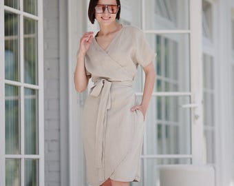May include: A woman wearing a beige linen wrap dress with a belt and brown sunglasses. She is standing in front of a white door with glass panels. She is wearing brown and white striped sandals.