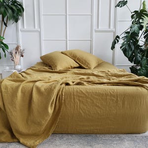 Op de afbeelding: Een bed met mosterdgele linnen beddengoed, inclusief een hoeslaken, dekbedovertrek en twee kussens. Het bed staat in een lichte kamer met witte muren en grote groene planten. Het beddengoed heeft een zachte, gekreukelde textuur.