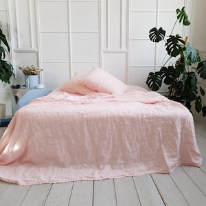 May include: A bed with light pink linen bedding, including a duvet cover and two pillows. The bed is in a bright room with white walls and a white wood floor. Two large potted plants are in the background.