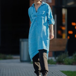 May include: A light blue linen tunic dress with a button-down front and long sleeves, worn with black trousers and sandals. The dress features an asymmetrical hemline. The model is standing on a paved walkway.
