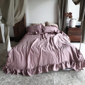 May include: A dusty rose-colored bedspread with ruffled edges and matching pillowcases. The bedding is on a bed with a canopy, creating a soft and inviting aesthetic. The room has a neutral color palette with wooden furniture and a vase of flowers.