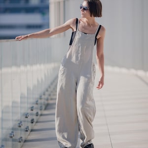 Könnte beinhalten: Eine Frau trägt einen hellbeigen Leinen-Jumpsuit mit einem schwarzen Tanktop darunter. Sie trägt eine schwarze Sonnenbrille und steht auf einer Betonfläche mit einem Glasgeländer im Hintergrund.
