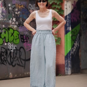 Puede incluir: Una mujer con un top blanco de tirantes y pantalones anchos de color azul claro con una cintura elástica blanca. Lleva zapatillas blancas y tiene las manos en las caderas. Está de pie frente a una pared con grafitis.