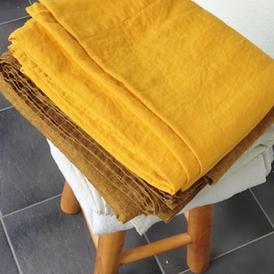May include: A stack of folded linen textiles in shades of yellow, brown, and white rests on a small wooden stool. The textiles appear to be sheets or blankets, with a textured, natural appearance. The stool has three legs.
