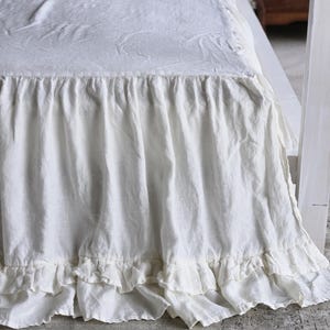 May include: A white bed skirt with a ruffled design. The skirt is made of a soft, textured fabric and hangs down to the floor. The bed is covered with a white sheet and pillows, creating a clean and inviting look.