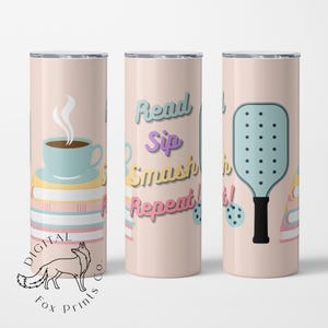 以下が含まれることがあります： ピンクとピーチ色のタンブラーで、コーヒーカップ、本の積み重ね、ピクルボールのパドル、そして「Read Sip Smash Repeat！」というテキストがデザインされています