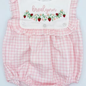May include: Pink and white gingham baby romper with a white bib featuring the name "Brealynn" embroidered in pink, along with strawberries and flowers. The romper has ruffled sleeves and trim, and snap closures.