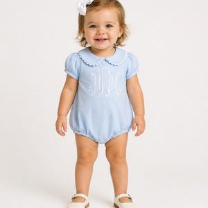 May include: A light blue baby romper with a white scalloped collar and puff sleeves. The romper features a white monogram and is paired with white shoes and a white bow. The baby is smiling.