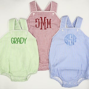 May include: Three baby rompers with different colored gingham patterns. The romper on the left is green and white, the middle romper is red and white, and the romper on the right is blue and white. Each romper has a different embroidered name on the front.