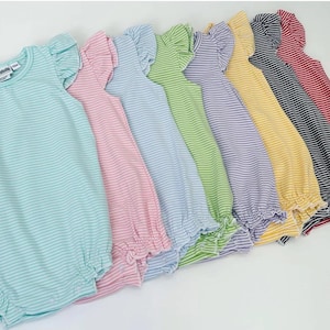 May include: A row of striped baby onesies in various colors, including light blue, pink, light purple, light green, yellow, black, and red. The onesies have ruffled sleeves and a gathered hem.