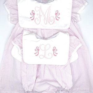 May include: Two pink and white striped dresses with white bibs and pink embroidered monograms. The larger dress has an "M" monogram and the smaller dress has an "L" monogram.