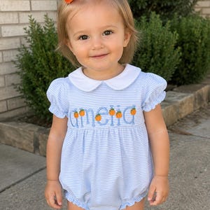 May include: A light blue and white striped bubble romper featuring a white Peter Pan collar, puff sleeves, and the name Amelia embroidered in blue script with small orange pumpkin accents between the letters, paired with a large orange hair bow.