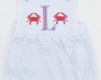 May include: Pale blue baby romper featuring red crab embroidery and the letter "L". The romper has white trim, button fastenings on the straps, and a gathered bottom. A charming outfit for a baby.