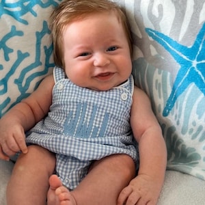 May include: A baby boy wearing a blue and white gingham romper with a blue embroidered monogram. The romper has a button closure at the neck and straps. The baby is lying on a white and blue patterned pillow.