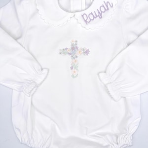 May include: White long-sleeved baby onesie with a scalloped collar, embroidered with a floral cross and the name "Rayah".