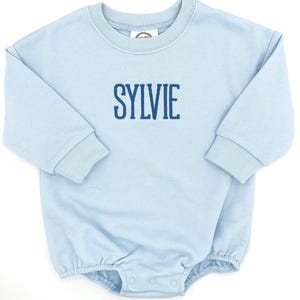 May include: A light blue long-sleeve romper featuring a ribbed crew neckline and ribbed cuffs. The name SYLVIE is embroidered in dark blue capital letters across the chest. The garment includes a snap-button closure at the bottom crotch area.