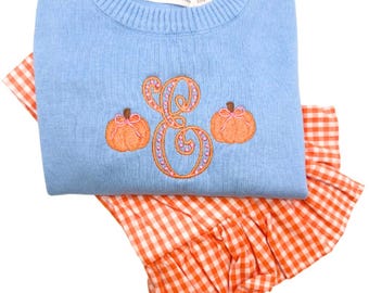 May include: A light blue sweater with a ribbed crew neck and the brand name "CAMPUR" on the label. The sweater features an embroidered orange letter "E" with pink accents and two orange pumpkins. It is paired with an orange and white gingham skirt.