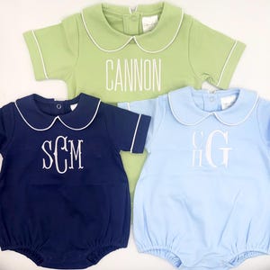 May include: Three baby rompers in green, navy blue, and light blue. Each romper has a white Peter Pan collar and trim. The green romper has the name "CANNON" embroidered. The navy blue romper has the initials "SCM". The light blue romper has the initials "CHG".