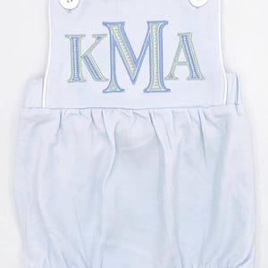 May include: A light blue baby onesie with white straps and white trim. The onesie has a large embroidered monogram of the letters "KMA" in blue, green, and white.