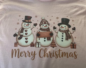 May include: A light pink t-shirt featuring a graphic of three snowmen wearing hats and scarves, surrounded by small gift boxes and snowflakes. The snowmen are positioned above the brown text that reads Merry Christmas.