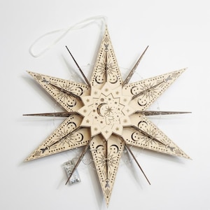 May include: A wooden star-shaped ornament with intricate celestial designs, including moons, stars, and an angel. The light-colored wood is laser-cut with dark brown details. A string is attached for hanging, and a string of lights is wrapped around the star.