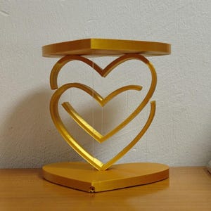 以下が含まれることがあります： ハート型のベースを持つ、金色の3次元ハートの彫刻。彫刻は、2つの絡み合ったハートで構成されています。