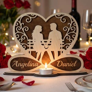 Può includere: Un portacandele a forma di cuore in legno con la silhouette di una coppia su una panchina. I nomi "Angelina" e "Daniel" sono incisi sulla base. Una candela a lumino accesa è al centro, con volute e ritagli decorativi.