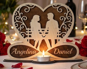 Può includere: Un portacandele a forma di cuore in legno con la silhouette di una coppia su una panchina. I nomi "Angelina" e "Daniel" sono incisi sulla base. Una candela a lumino accesa è al centro, con volute e ritagli decorativi.