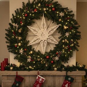 May include: A large Christmas wreath with a white star in the center, decorated with red and gold ornaments, pinecones, and lights. Below, a stone mantel displays three Christmas stockings.