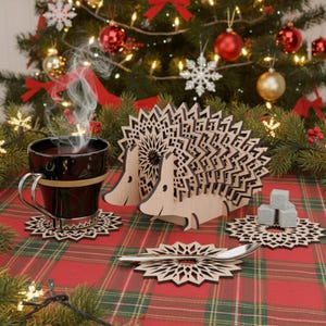 Może przedstawiać: Świąteczna scena z czarnym kubkiem z parą, drewnianymi podstawkami w kształcie jeża i podstawkami w kształcie płatków śniegu. Podstawki i uchwyty wykonane są z jasnego drewna. Tło obejmuje udekorowaną choinkę i czerwoną i zieloną kratowaną serwetę.