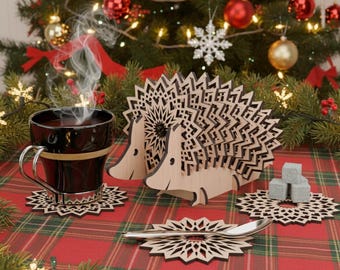 May include: A Christmas-themed image showing a black mug with steam, wooden hedgehog-shaped coaster holders, and snowflake coasters. The coasters and holders are made of light-coloured wood. The background includes a decorated Christmas tree and a red and green plaid tablecloth.