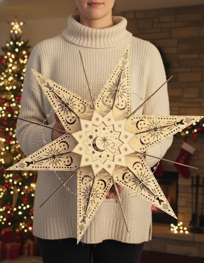 May include: A large, intricately designed wooden star decoration. The star features cut-out patterns of moons, stars, and celestial designs. It is held by a person wearing a cream-colored sweater, with a Christmas tree and fireplace in the background.