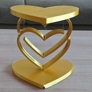 以下が含まれることがあります： 中空の中心を持つ、金色の3次元ハートの彫刻。彫刻は、互いに絡み合った2つのハートでできており、1つはもう1つの上にあります。